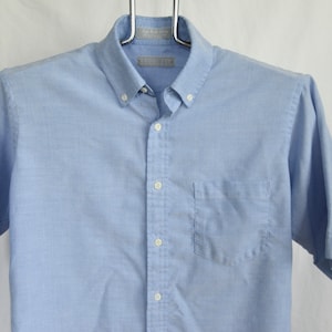 Puede incluir: Una camisa de manga corta de color azul claro con un bolsillo en el pecho. La camisa tiene un cuello blanco y botones blancos.