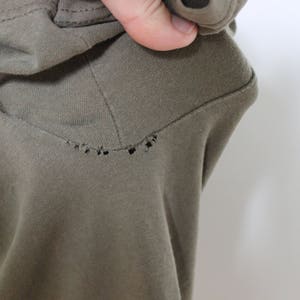 Op de afbeelding: Een close-up van een groen T-shirt met een klein gaatje in de stof. Het T-shirt is gemaakt van een zacht, comfortabel materiaal.