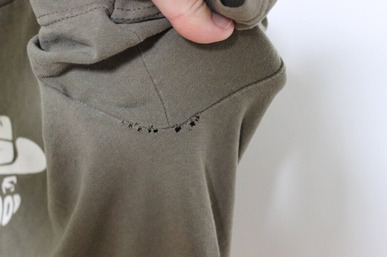 Op de afbeelding: Een close-up van een groen T-shirt met een klein gaatje in de stof. Het T-shirt is gemaakt van een zacht, comfortabel materiaal.
