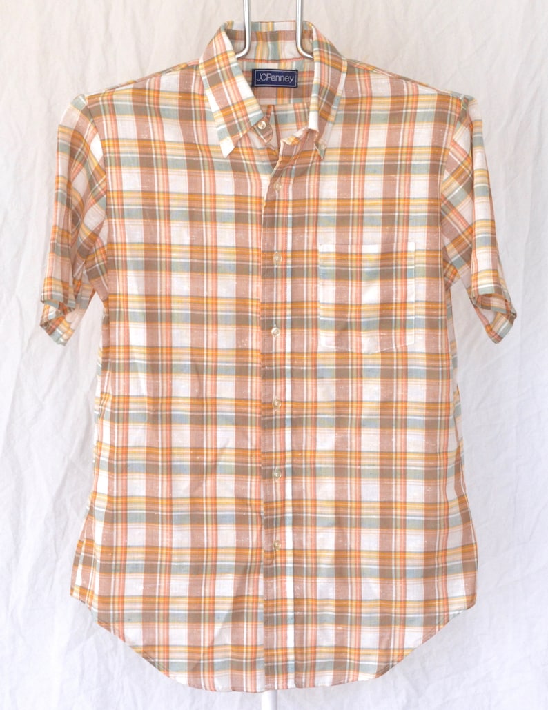 May include: A short-sleeved button-down shirt with a plaid pattern in shades of orange, brown, and white. The shirt has a pocket on the left chest and a label that reads "JCPenney".