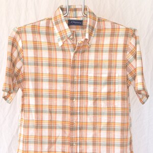 May include: A short-sleeved button-down shirt with a plaid pattern in shades of orange, brown, and white. The shirt has a pocket on the left side and a label that reads "JCPenney".