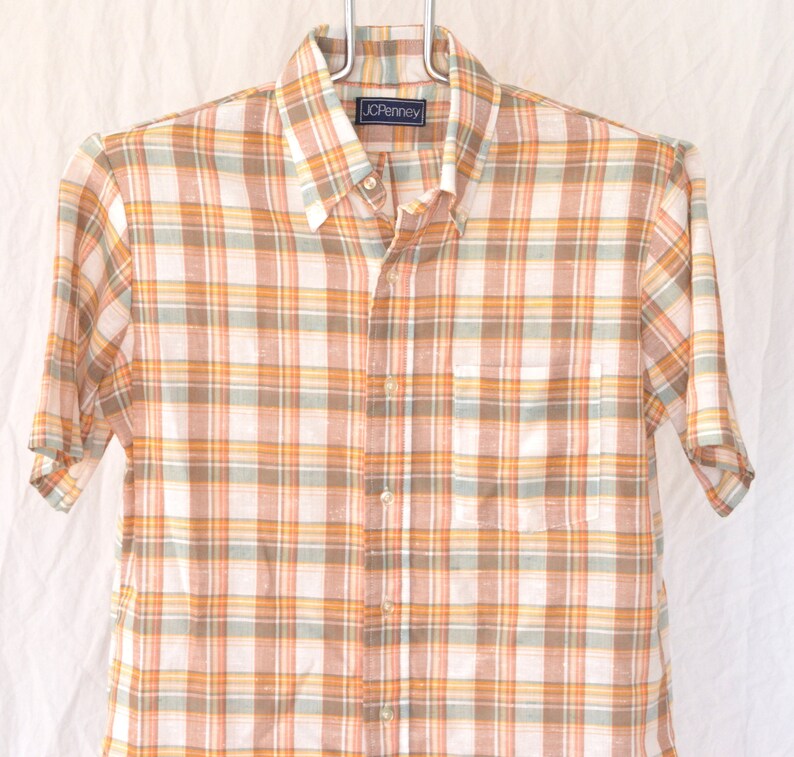 May include: A short-sleeved button-down shirt with a plaid pattern in shades of orange, brown, and white. The shirt has a pocket on the left side and a label that reads "JCPenney".