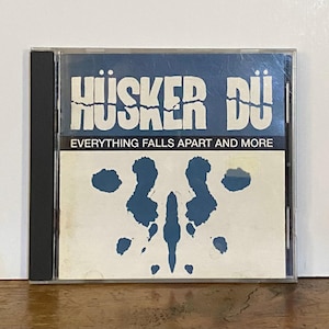 Op de afbeelding: Een cd-hoesje met de band Hüsker Dü. De bovenste helft van de hoes is blauw met de naam van de band in witte letters. De onderste helft is wit met een blauw inktvlekkenontwerp. De tekst "EVERYTHING FALLS APART AND MORE" is gedrukt.