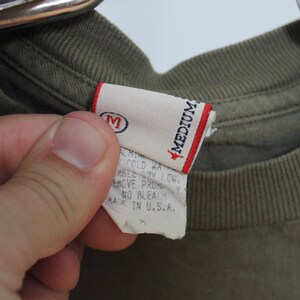 Op de afbeelding: Een groen T-shirt met een wit label waarop "Medium" staat en wasinstructies. Het label vermeldt ook "Made in U.S.A."