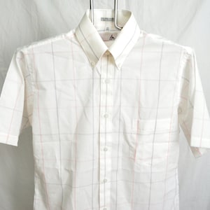 May include: A white short-sleeved button-down shirt with a subtle plaid pattern in light pink and gray. The shirt has a button-down collar and a single chest pocket. The shirt is hanging on a metal hanger.