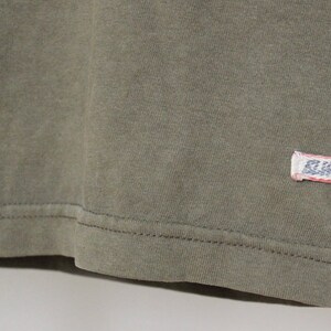 Op de afbeelding: Een close-up van de zoom van een groen T-shirt met een klein wit label met de tekst "R.L.P.D."