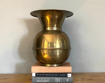 May include: A gold metal vase with a wide, flared top and a round body. The vase is sitting on top of three books, including "Blood Meridian" by Cormac McCarthy and "To Kill a Mockingbird" by Harper Lee.