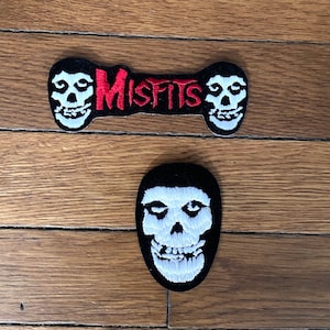 Könnte beinhalten: Zwei gestickte Patches in Schwarz und Weiß mit dem Logo der Band Misfits. Das Logo ist ein Schädel mit schwarzem Hintergrund und weißen Augen und Zähnen. Das Wort "Misfits" ist in roten Buchstaben geschrieben. Die Patches befinden sich auf einer Holzoberfläche.