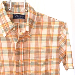 May include: A short-sleeved button-down shirt with a plaid pattern in shades of orange, brown, and white. The shirt has a pocket on the left side and a label that reads "JCPenney".
