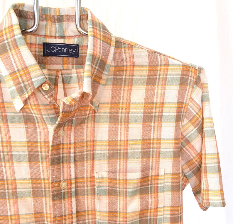 May include: A short-sleeved button-down shirt with a plaid pattern in shades of orange, brown, and white. The shirt has a pocket on the left side and a label that reads "JCPenney".