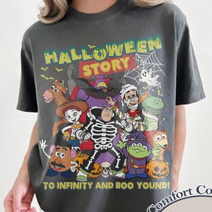 May include: Dark gray Comfort Colors t-shirt featuring a Halloween-themed graphic with the text "HALLOWEEN STORY" and "TO INFINITY AND BOO YOUND!" The graphic includes cartoon characters and pumpkins. The shirt has a crew neck.