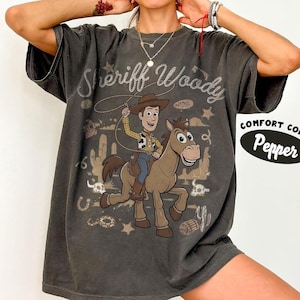 May include: A charcoal gray short-sleeve t-shirt featuring a graphic of a cowboy character riding a horse with a lasso. The text Sheriff Woody is printed above the character. A Comfort Colors Pepper label is visible on the right side of the shirt.