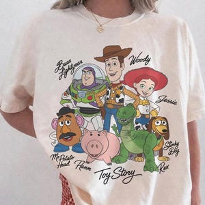 May include: Ivory-coloured t-shirt featuring a graphic of Toy Story characters including Woody, Buzz Lightyear, and Jessie. The shirt has the words "Toy Story" printed below the characters. The shirt is labelled "Comfort Colors" and "Ivory".