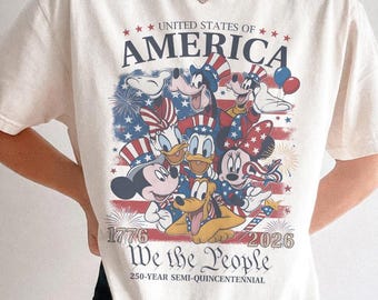 以下が含まれることがあります： アイボリー色のTシャツ。愛国的なデザインで、「UNITED STATES OF AMERICA」と「We the People」の文字がプリントされています。漫画のキャラクターと1776年と2026年の年号が描かれており、250周年を祝っています。
