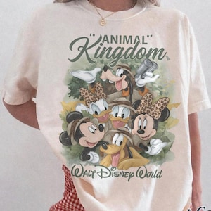 May include: Ivory-colored t-shirt featuring a graphic of Disney characters, including Mickey and Minnie Mouse, Donald Duck, and Goofy, with the text "Animal Kingdom" and "Walt Disney World". The shirt is a Comfort Colors brand.