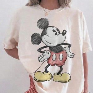 May include: Ivory-colored t-shirt featuring a sketch of Mickey Mouse in black, red, and yellow. The shirt has a relaxed fit and is paired with red and white checkered shorts. The text "Comfort Colors Ivory" is printed on the bottom right.