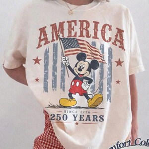 Pu&ograve; includere: T-shirt color avorio con grafica di Mickey Mouse con bandiera americana. La parola "AMERICA" &egrave; stampata sopra la grafica, con "250 YEARS" sotto. La maglietta &egrave; del marchio Comfort Colors.