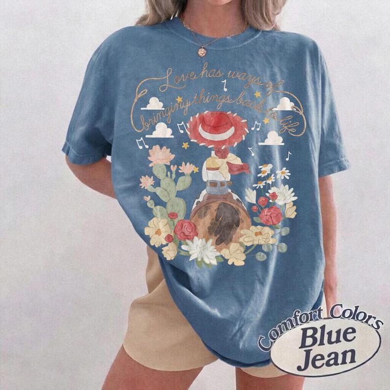 Peut inclure: Un t-shirt bleu avec un motif repr&eacute;sentant une cow-girl au chapeau rouge &agrave; cheval, entour&eacute;e de cactus, de fleurs, de nuages et de notes de musique. Le texte indique "Love has ways of bringing things back to life". Col rond et manches courtes.