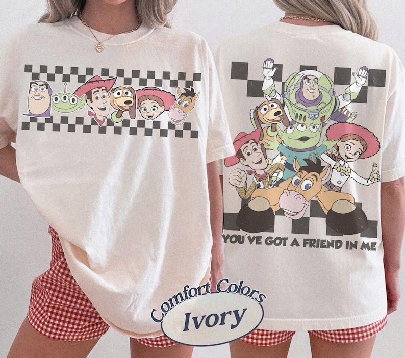 May include: Ivory-colored t-shirt featuring cartoon characters from a popular animated film. The front displays a checkered pattern with character faces. The back of the shirt shows a group of characters with the text "YOU'VE GOT A FRIEND IN ME."