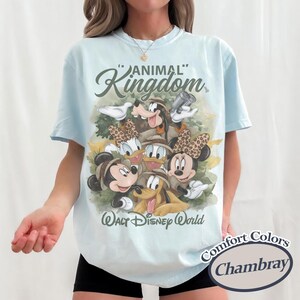 May include: Light blue t-shirt featuring a graphic of Disney characters, including Mickey and Minnie Mouse, Donald and Daisy Duck, Goofy, and Pluto. The shirt has the text "ANIMAL KINGDOM" and "Walt Disney World". The shirt is a Comfort Colors Chambray.