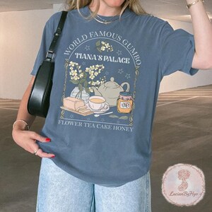 May include: A blue t-shirt with a graphic design featuring the text "WORLD FAMOUS GUMBO" and "TIANA'S PALACE". The design includes illustrations of a teapot, honey jar, teacup, cake, and flowers. The words "FLOWER TEA CAKE HONEY" are also printed on the shirt.
