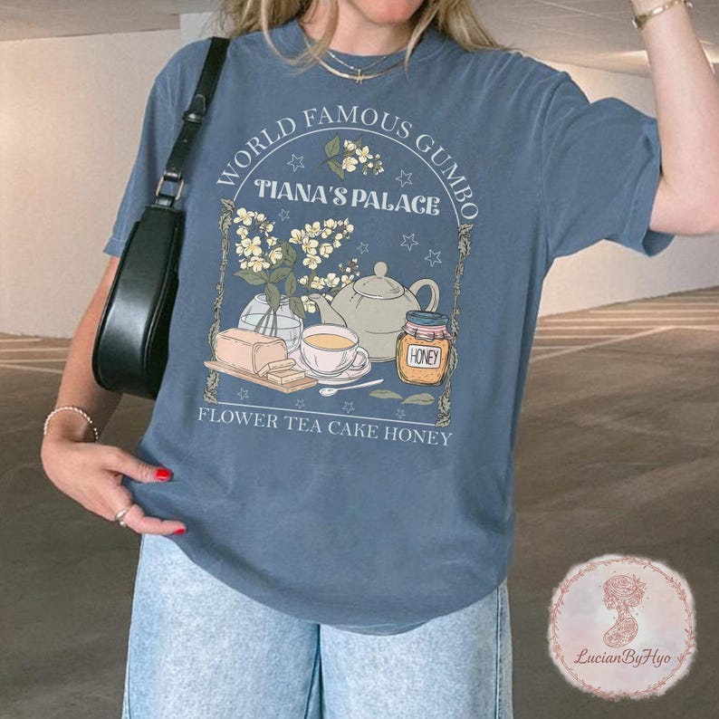 May include: A blue t-shirt with a graphic design featuring the text "WORLD FAMOUS GUMBO" and "TIANA'S PALACE". The design includes illustrations of a teapot, honey jar, teacup, cake, and flowers. The words "FLOWER TEA CAKE HONEY" are also printed on the shirt.