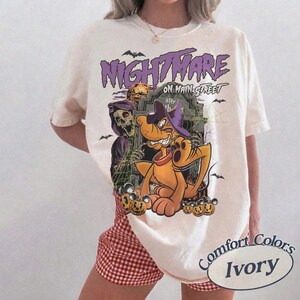 May include: Ivory-colored t-shirt with a Halloween-themed graphic. The design features a cartoon dog wearing a witch hat, a skeleton, and the words "Nightmare on Main Street." Bats and pumpkins are also included in the design.