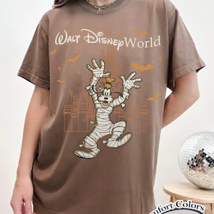 以下が含まれることがあります： エスプレッソ色のTシャツ。ハロウィーンをテーマにした、ミイラに扮したグーフィーのグラフィックと「ウォルト・ディズニー・ワールド」の文字がプリントされています。デザインにはコウモリと城のシルエットが含まれています。Comfort Colorsのシャツです。