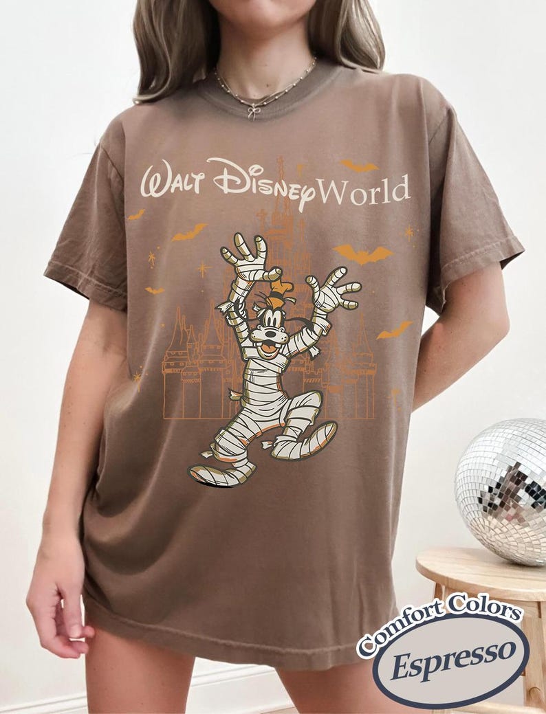 以下が含まれることがあります： エスプレッソ色のTシャツ。ハロウィーンをテーマにした、ミイラに扮したグーフィーのグラフィックと「ウォルト・ディズニー・ワールド」の文字がプリントされています。デザインにはコウモリと城のシルエットが含まれています。Comfort Colorsのシャツです。