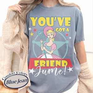 Op de afbeelding: Blauw Comfort Colors T-shirt in jeanskleur met een stripfiguur met een herdersstaf. De tekst luidt "YOU'VE GOT A FRIEND Jume!" in geel en wit. Het personage draagt een roze jurk. Het T-shirt is blauw.