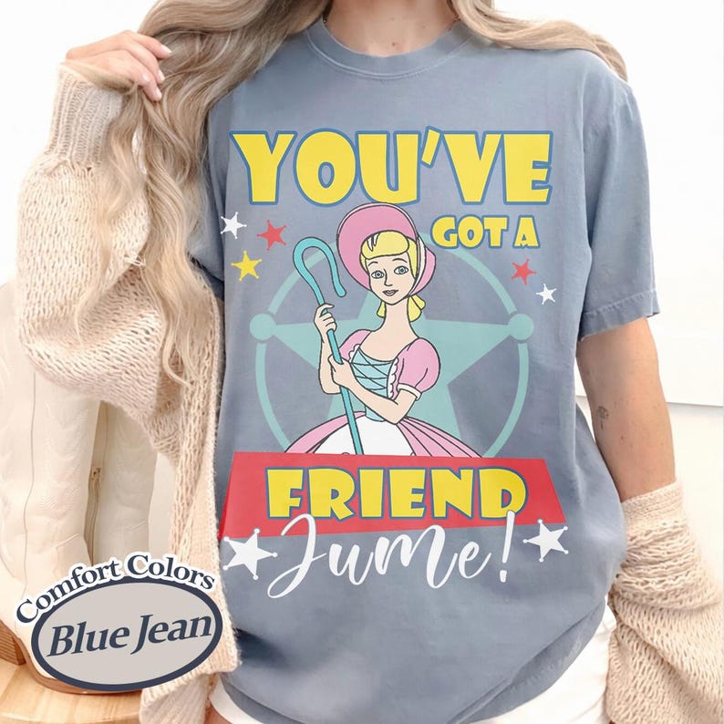 Op de afbeelding: Blauw Comfort Colors T-shirt in jeanskleur met een stripfiguur met een herdersstaf. De tekst luidt "YOU'VE GOT A FRIEND Jume!" in geel en wit. Het personage draagt een roze jurk. Het T-shirt is blauw.