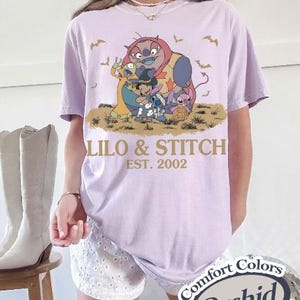 K&ouml;nnte beinhalten: Ein helllila T-Shirt mit einer Grafik von Lilo & Stitch-Charakteren und dem Text "Lilo & Stitch EST. 2002". Das Design umfasst Stitch, Lilo und andere Charaktere. Das Shirt ist ein Comfort Colors Orchid.