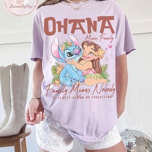 May include: A light purple t-shirt featuring the words "OHANA Means Family" in brown lettering, with an illustration of Stitch and Lilo hugging. The text "Family Means Nobody Gets Left Behind Or Forgotten" is below the illustration.