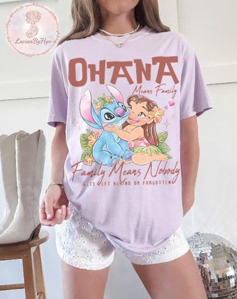 May include: A light purple t-shirt featuring the words "OHANA Means Family" in brown lettering, with an illustration of Stitch and Lilo hugging. The text "Family Means Nobody Gets Left Behind Or Forgotten" is below the illustration.