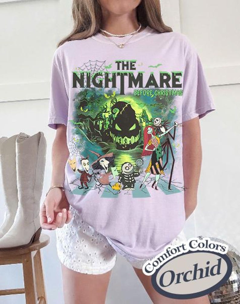 K&ouml;nnte beinhalten: Ein helllila T-Shirt mit einer Grafik aus "The Nightmare Before Christmas". Das Design zeigt Jack Skellington und Sally, mit dem Filmtitel in Gr&uuml;n und Schwarz. Das Shirt ist mit "Comfort Colors Orchid" gekennzeichnet.