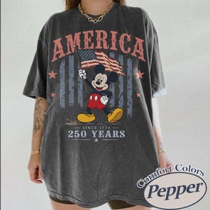 Pu&ograve; includere: T-shirt grigio scuro con Topolino che tiene una bandiera americana. La parola "AMERICA" &egrave; stampata in alto, con "SINCE 1776 - 250 YEARS" sotto. La maglietta &egrave; di Comfort Colors, Pepper.