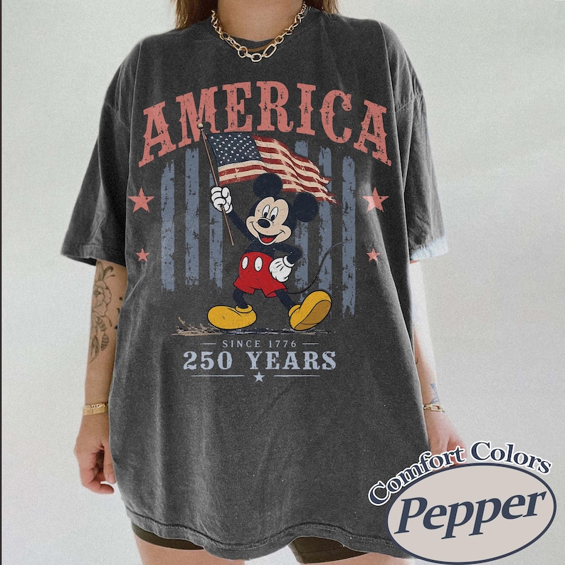 Pu&ograve; includere: T-shirt grigio scuro con Topolino che tiene una bandiera americana. La parola "AMERICA" &egrave; stampata in alto, con "SINCE 1776 - 250 YEARS" sotto. La maglietta &egrave; di Comfort Colors, Pepper.