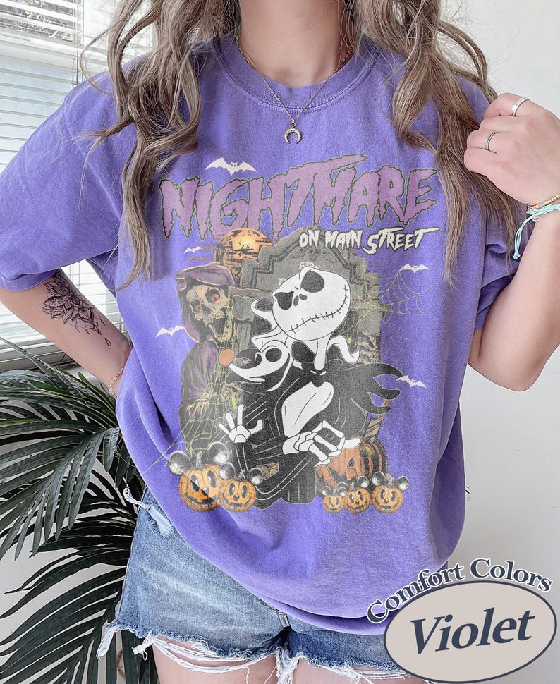 May include: Violet Comfort Colors t-shirt featuring a graphic from "The Nightmare Before Christmas." The design includes Jack Skellington, Zero, and pumpkins. The text "NIGHTMARE ON MAIN STREET" is printed above the characters.