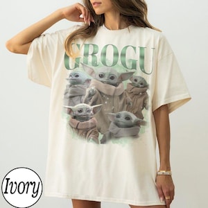 Puede incluir: Una camiseta de manga corta color marfil con un gran estampado gr&aacute;fico de cinco peque&ntilde;as criaturas verdes con orejas y ojos grandes, vestidas con t&uacute;nicas color canela. El texto GROGU est&aacute; impreso en letras verdes grandes sobre ellas.