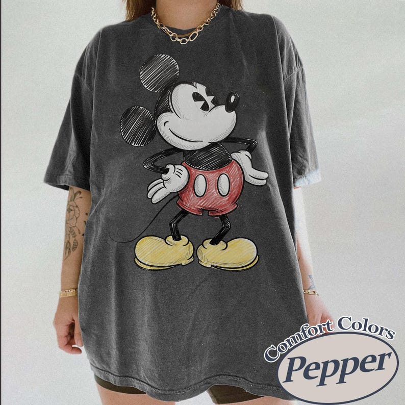 May include: Dark gray t-shirt featuring a sketched Mickey Mouse graphic. Mickey is depicted with a white face, black ears, red shorts, and yellow shoes. The shirt has the text "Comfort Colors Pepper" in a white oval.