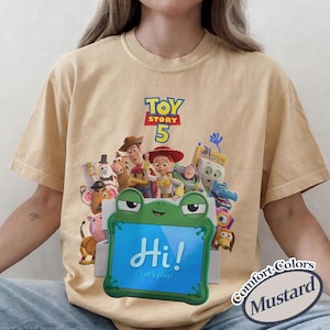 May include: A mustard yellow crew neck t-shirt featuring a graphic print of Toy Story 5 characters including Woody, Jessie, and Buzz Lightyear. In the foreground, a green frog-shaped tablet displays the text Hi! Let's play! on its screen.