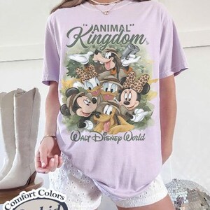 May include: A light purple t-shirt featuring a graphic of Disney characters, including Mickey and Minnie Mouse, Goofy, Donald and Daisy Duck, and Pluto. The shirt has the words "Animal Kingdom" and "Walt Disney World" printed on it.