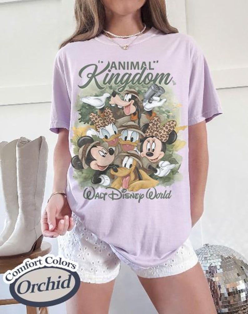 May include: A light purple t-shirt featuring a graphic of Disney characters, including Mickey and Minnie Mouse, Goofy, Donald and Daisy Duck, and Pluto. The shirt has the words "Animal Kingdom" and "Walt Disney World" printed on it.