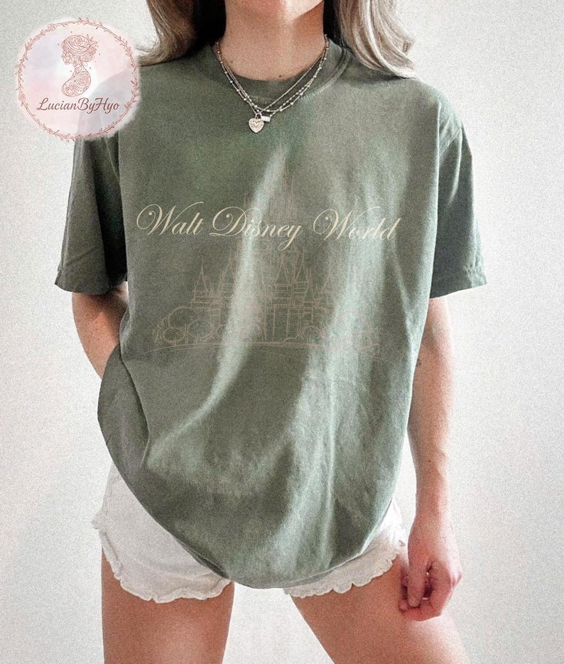 May include: Olive green t-shirt with the text "Walt Disney World" in a cursive font, and a line drawing of a castle. The shirt is a relaxed fit and has short sleeves. The shirt is paired with white shorts.
