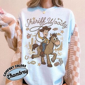 May include: A light blue crewneck sweatshirt featuring a graphic of Sheriff Woody riding a brown horse. The design includes the text Sheriff Woody, a lasso, cacti, stars, and desert elements in brown ink. A Comfort Colors Chambray label is visible.