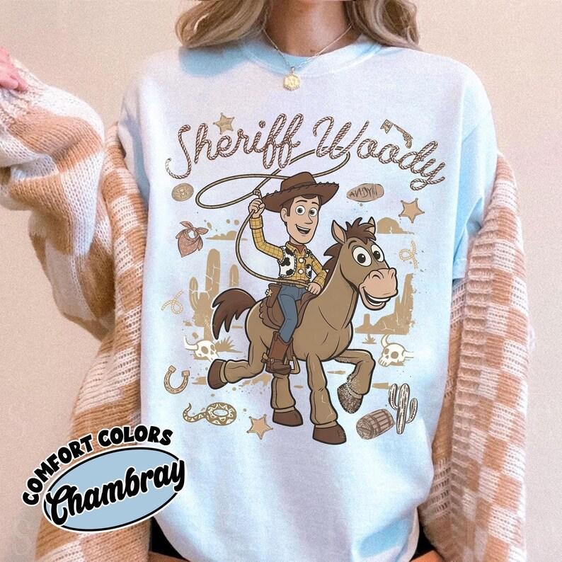 May include: A light blue crewneck sweatshirt featuring a graphic of Sheriff Woody riding a brown horse. The design includes the text Sheriff Woody, a lasso, cacti, stars, and desert elements in brown ink. A Comfort Colors Chambray label is visible.