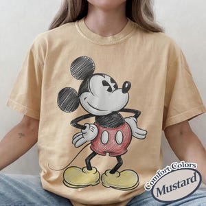 May include: Mustard-colored t-shirt featuring a sketch of Mickey Mouse in black, white, and red. The cartoon character is in a standing pose with his hands on his hips. The text "Comfort Colors Mustard" is in a white oval on the bottom right.