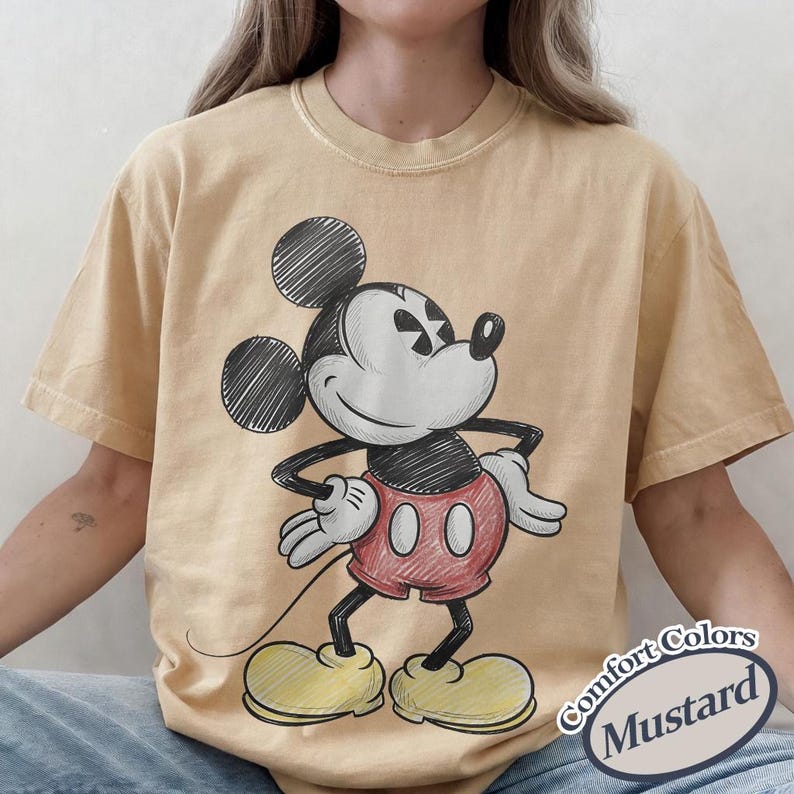 May include: Mustard-colored t-shirt featuring a sketch of Mickey Mouse in black, white, and red. The cartoon character is in a standing pose with his hands on his hips. The text "Comfort Colors Mustard" is in a white oval on the bottom right.