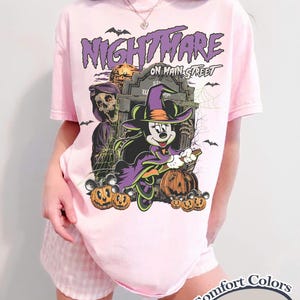 May include: A light pink t-shirt with a Halloween-themed graphic. The graphic features the text "Nightmare on Main Street" above an illustration of Mickey Mouse in a witch costume, pumpkins, and a graveyard scene. The shirt is labeled "Comfort Colors Blossom".