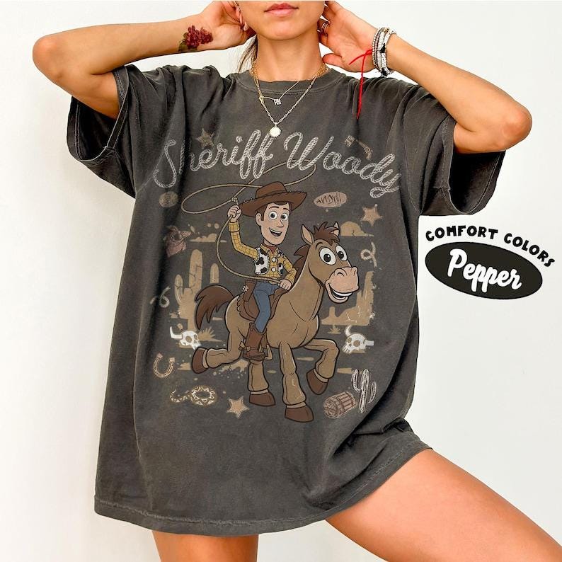 May include: A charcoal gray short-sleeve t-shirt featuring a graphic of a cowboy character riding a horse with a lasso. The text Sheriff Woody is printed above the character. A Comfort Colors Pepper label is visible on the right side of the shirt.